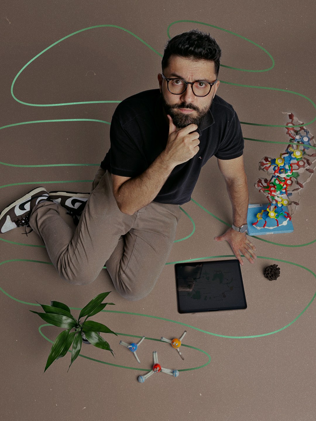 O professor Vitor Hugo, visto de cima, cercado por um modelo de dupla-hélice de DNA, modelos moleculares, tablet e planta